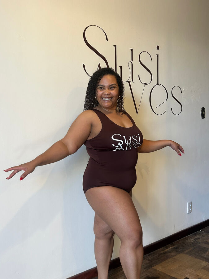 Modelo sorrindo com collant marrom Susi Alves em frente ao logo do estúdio